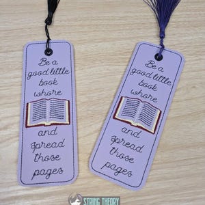 Op de afbeelding: Twee lavendelkleurige bladwijzers met zwarte en paarse kwastjes. Elke bladwijzer heeft de geborduurde tekst "Be a good little book whore and spread those pages" met een geborduurde boekillustratie.