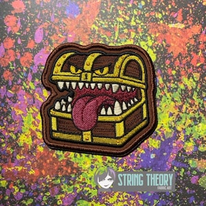 May include: An embroidered patch featuring a brown treasure chest with gold trim, sharp white teeth, and a large pink tongue hanging out. The chest has yellow eyes and a dark brown border, designed as a mimic creature with a menacing expression.