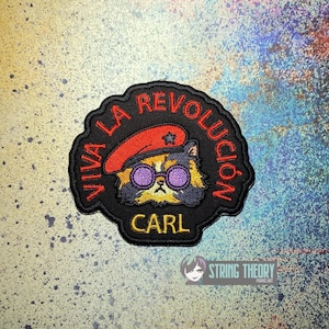 May include: A black embroidered patch featuring a calico cat wearing a red beret with a star and purple round glasses. The text VIVA LA REVOLUCIÓN is arched in red thread above the cat, and the name CARL is embroidered in yellow below the cat.