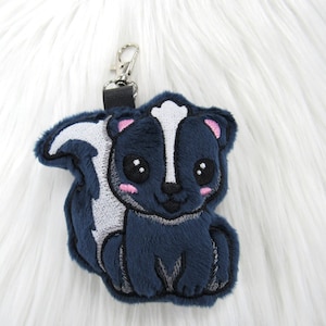 May include: A plush blue and white skunk keychain with a black nose and big eyes. The keychain has a silver metal ring.