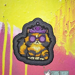May include: An embroidered patch featuring a fluffy calico cat with orange, black, and white fur. The cat wears a purple tiara and round purple-rimmed glasses. The patch has a black border and is set against a vibrant yellow and pink background.