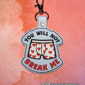May include: A light blue keychain featuring an embroidered graphic of white boxer shorts with red hearts. The text YOU WILL NOT is embroidered in black above the shorts, and BREAK ME is embroidered in red below. A silver metal lobster clasp is attached.