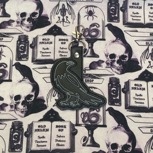 May include: A black raven charm with a gold key ring. The charm is on a background of a white fabric with a repeating pattern of skulls, ravens, and jars with the text "Old Salem" and "Book of" on them. The text "House of Salem's Old Curiosity Shop" is also visible.