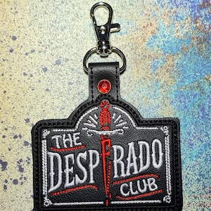 May include: A black faux leather keychain featuring an embroidered design with the text THE DESPERADO CLUB in white and red. A vertical red dagger graphic sits between the words. A single red rhinestone is set above the text, and a silver metal lobster clasp is attached to the top.