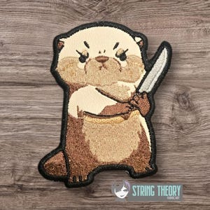 May include: Embroidered patch featuring a cartoon otter holding a knife. The otter has a stern expression and is rendered in shades of brown, beige, and black. The patch has a black border and the text "STRING THEORY FABRIC ART" is visible.