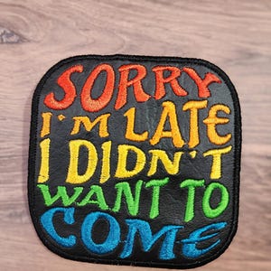 Puede incluir: Parche cuadrado negro con la frase bordada "SORRY I'M LATE I DIDN'T WANT TO COME" en colores arcoíris. El parche tiene un borde negro y probablemente está hecho de tela. El logotipo de String Theory Fabric Art está en la esquina inferior derecha.