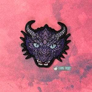 May include: Embroidered dragon patch featuring a detailed purple and black design. The dragon has blue eyes, horns, and sharp teeth. The patch is on a pink background. The String Theory logo is visible.
