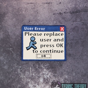 Op de afbeelding: Een blauwe en witte geborduurde patch met een computerfoutmelding die zegt "Gebruikersfout" en "Vervang de gebruiker en druk op OK om door te gaan". De patch heeft een blauwe rand en een rode X in de rechterbovenhoek. Er is een blauw stickfiguurpictogram in het midden van de patch.