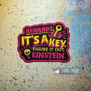 May include: A rectangular embroidered patch with a black background and a bright pink border. The text reads REWARD IT'S A KEY. FIGURE IT OUT, EINSTEIN. It features a yellow key icon and a yellow lightbulb icon with a skull inside the bulb.