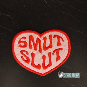 May include: A heart-shaped patch with a white center and a red border. The words "SMUT SLUT" are embroidered in red across the center. The patch is likely for clothing or accessories.