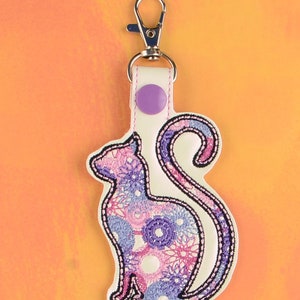 May include: A white leather keychain with a purple button and a pink and purple embroidered cat design. The keychain has a silver metal clasp.