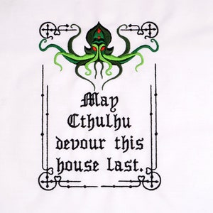 Pode incluir: Tecido branco com um desenho bordado. Apresenta uma criatura verde semelhante a Cthulhu com olhos vermelhos e tentáculos. O texto diz "May Cthulhu devour this house last." O desenho é emoldurado com elementos decorativos pretos.