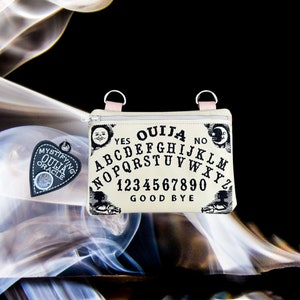 May include: A beige zippered pouch with a black and white Ouija board design. The pouch has two pink zipper pulls and the text "Ouija" is printed in black at the top. The pouch is sitting on a white and gray smoke-like background.