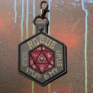May include: A hexagonal gray keychain featuring an embroidered red twenty-sided die with a skull icon in the center. The text ROGUE YOUR STUFF IS MY STUFF is stitched in gold thread around the die. It includes a black metal lobster claw clasp.