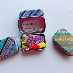 May include: Three small, rectangular tins with colorful patterned tape designs. One tin is open and contains a small red and yellow object, a few pieces of patterned paper, and a note that says "Say a "little" something."
