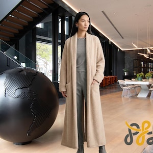 May include: A woman wearing a long, beige cardigan with pockets, paired with gray pants and a top. The background features a black globe sculpture and a modern interior with a staircase and a dining area.