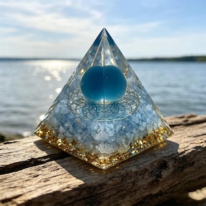 May include: A clear resin pyramid containing a central blue sphere, a gold-coloured geometric flower of life symbol, a layer of light blue stone chips, and a base layer of gold-coloured metallic flakes, creating a multi-layered decorative object.