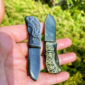 Puede incluir: Dos cuchillos de cristal de obsidiana con diseños intrincados. Los cuchillos son negros con acentos iridiscentes verdes y dorados. Los cuchillos se sostienen en una mano.