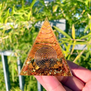May include: A clear pyramid with a copper wire spiral and various crystals inside. The pyramid is made of resin and has a golden color.