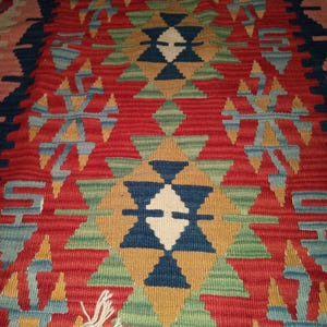 May include: A vibrant, hand-woven rug with a geometric design. The rug features a central pattern of red, green, blue, and gold shapes, with a white diamond in the center. The edges are trimmed with tassels. The rug is a traditional style.