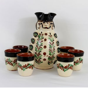 May include: A ceramic pitcher and matching cups set. The pitcher is off-white with a black top and a face design. The cups are off-white with a black rim and a brown and green leaf design. The set is likely handmade.