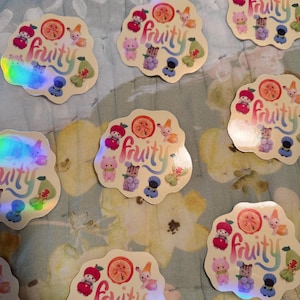 May include: A collection of holographic stickers with a whimsical design. Each sticker features the word "fruity" in a colorful script, surrounded by cartoon fruit and adorable characters. The stickers have a pearlescent, iridescent finish.
