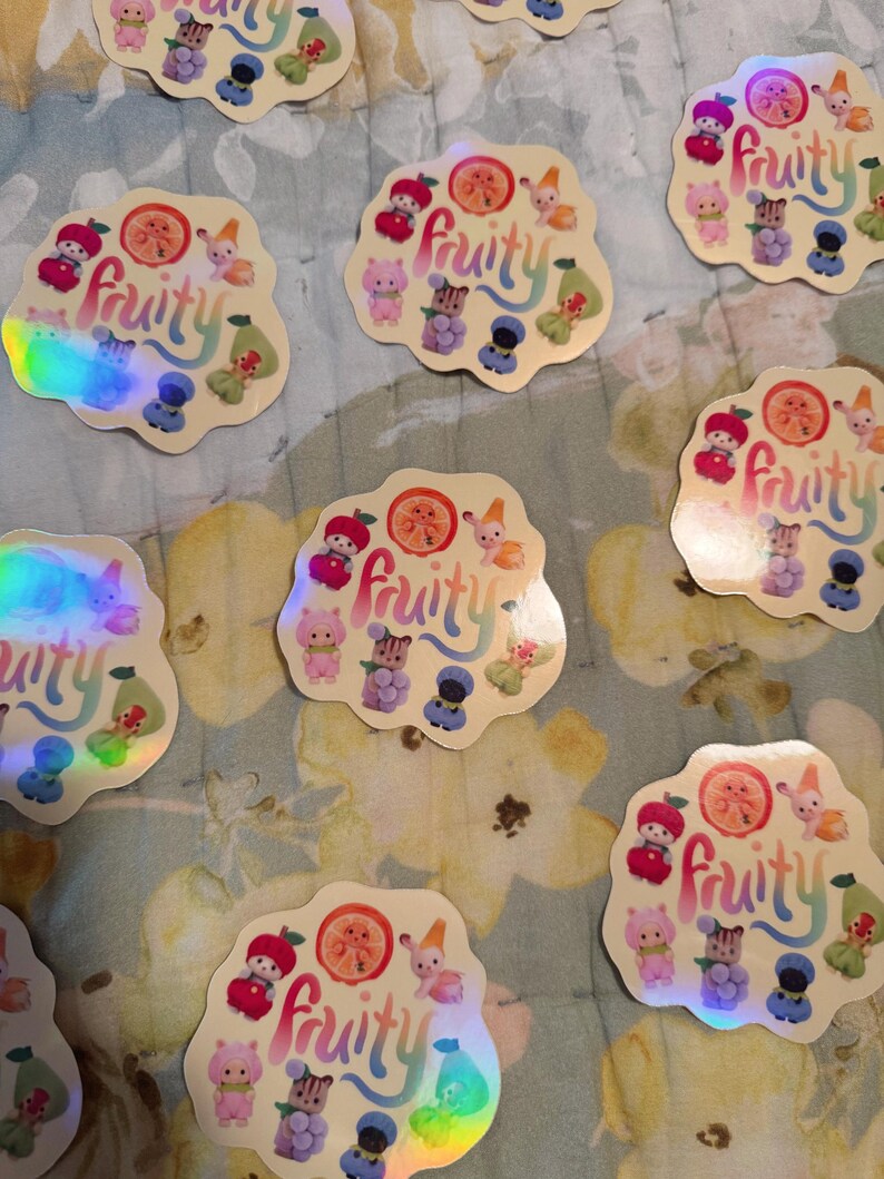 May include: A collection of holographic stickers with a whimsical design. Each sticker features the word "fruity" in a colorful script, surrounded by cartoon fruit and adorable characters. The stickers have a pearlescent, iridescent finish.