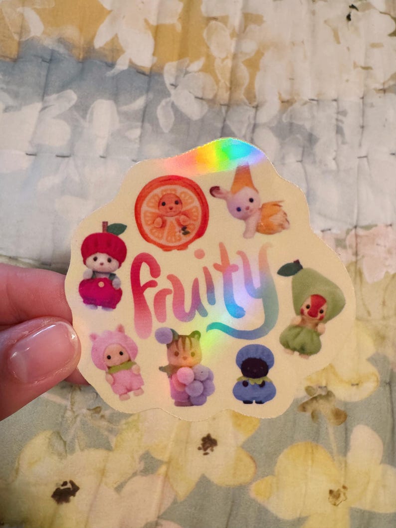 May include: A holographic sticker with the word "Fruity" in rainbow colors. The sticker features cartoon characters designed as fruits, including an apple, orange slice, pear, and other whimsical figures. The sticker is held by a hand.