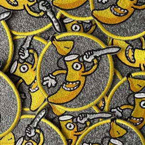 Puede incluir: Una colección de parches bordados amarillos y grises con un personaje de plátano de dibujos animados que sostiene un cuchillo. El personaje de plátano tiene una sonrisa de dientes y un solo ojo.