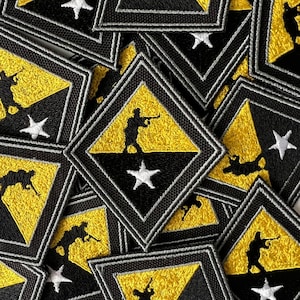 Puede incluir: Primer plano de varios parches bordados con un diseño amarillo y negro con una estrella blanca. Los parches tienen forma de diamante y presentan la silueta de una persona sosteniendo un rifle.