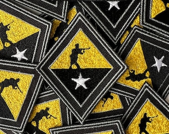 Puede incluir: Primer plano de varios parches bordados con un diseño amarillo y negro con una estrella blanca. Los parches tienen forma de diamante y presentan la silueta de una persona sosteniendo un rifle.
