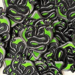 Puede incluir: Primer plano de varios parches de hierro bordados en verde y negro con un diseño de serpiente. Los parches están dispuestos en un patrón aleatorio.