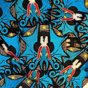 Puede incluir: Primer plano de varios parches bordados en azul y dorado que representan un pulpo estilizado con alicates para tentáculos. El pulpo está rodeado por un borde en forma de estrella.