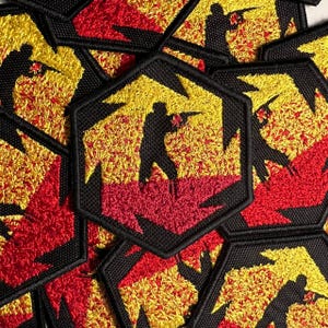 Puede incluir: Un conjunto de parches bordados negros, rojos y amarillos con la silueta de una persona sosteniendo un arma. Los parches son hexagonales y tienen una textura peluda.
