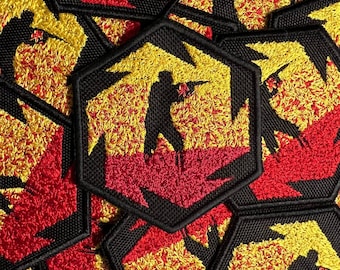 Puede incluir: Un conjunto de parches bordados negros, rojos y amarillos con la silueta de una persona sosteniendo un arma. Los parches son hexagonales y tienen una textura peluda.
