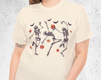 May include: Cream-coloured t-shirt with a graphic of three dancing skeletons, a pumpkin, bats, and autumn leaves. The word "Natural" is printed in the upper left corner. The design is in shades of brown and orange.