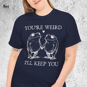 May include: Navy blue t-shirt with a white graphic of two frogs holding hands, with the text "YOU'RE WEIRD I'LL KEEP YOU" above and below the frogs. A small white heart is between the words "WEIRD" and "I'LL".