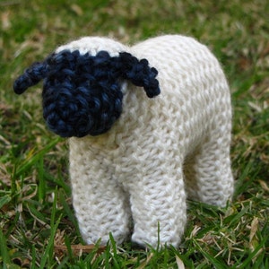 May include: A white knitted sheep with a black face and ears. The sheep is standing on a bed of green grass.