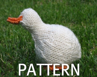 May include: A white knitted goose with an orange beak sitting on green grass. The goose is a handmade knitted toy.