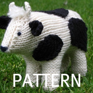 May include: A white knitted cow with black spots, standing on green grass. The cow has a white face with black eyes and a black nose. The word "PATTERN" is printed in white letters on the grass in front of the cow.