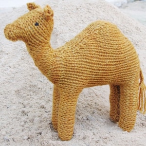 May include: A knitted camel toy made of brown yarn. The camel is standing on a sandy surface.