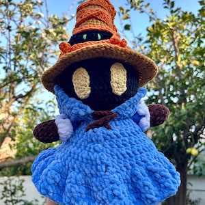 Puede incluir: Muñeca de ganchillo hecha a mano de un personaje mago, con cabeza y cuerpo negros, que lleva una túnica azul, un sombrero marrón y guantes. La muñeca tiene ojos beige y se sostiene frente a un fondo de follaje verde y cielo azul.