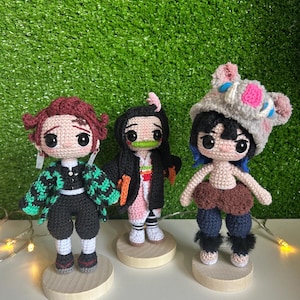 May include: Three crocheted dolls on wooden bases. The dolls are dressed in detailed outfits, including a green and black patterned jacket, a pink kimono, and a boar-themed hat. The dolls have detailed hairstyles and facial features.