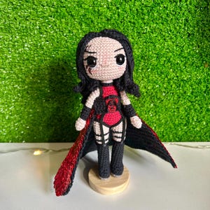 May include: A handmade crochet doll with black hair and a red and black outfit. The doll has a black cape with red accents and is standing on a small wooden base. The doll's face has black eyes and a small scar.