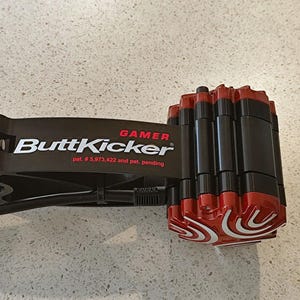May include: Black and red ButtKicker Gamer, a device for enhancing gaming experiences. The device features a black frame with red accents and the text "ButtKicker Gamer". It has a rotating red knob and a series of red and black cylinders.