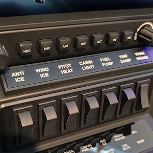 May include: Close-up of a black aircraft control panel with various switches, knobs, and labeled buttons. Text includes "ANTI ICE", "WIND ICE", "PITOT HEAT", "CABIN LIGHT", "FUEL PUMP", "MASTER WARNING", and "ENGINE FIRE".