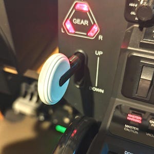 May include: A black flight simulator gear lever featuring a white circular knob, positioned next to a triangular indicator light labeled GEAR with N, L, and R markings. The panel includes UP and DOWN text labels, a MASTER WARNING light, and a NOSE DOWN control.