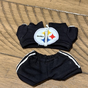 May include: Black doll-sized athletic outfit featuring a jacket and shorts. The jacket has a white zipper and a Steelers logo patch. The shorts have white stripes down the sides. The outfit is made of a soft, dark fabric.