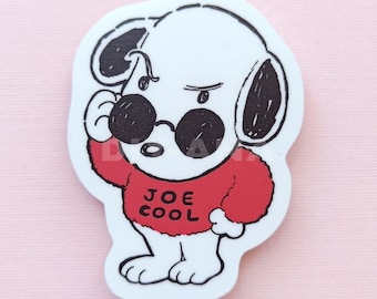 May include: A white cartoon dog with black ears and sunglasses, wearing a red sweater with the text "JOE COOL". The dog is giving a side-eye expression, with one paw raised. The design is outlined in black.
