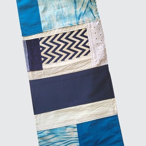 May include: A long, rectangular patchwork table runner with a white background and blue, beige, and white patterned fabric squares. The runner is made of different fabrics with various textures and patterns.